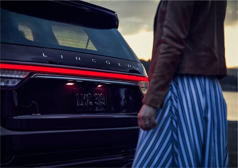 A person is shown near the rear of a 2023 Lincoln Aviator® SUV as the Lincoln Embrace illuminates the rear lights | Beach Lincoln in Myrtle Beach SC