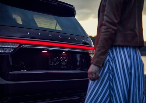 A person is shown near the rear of a 2024 Lincoln Aviator® SUV as the Lincoln Embrace illuminates the rear lights | Beach Lincoln in Myrtle Beach SC